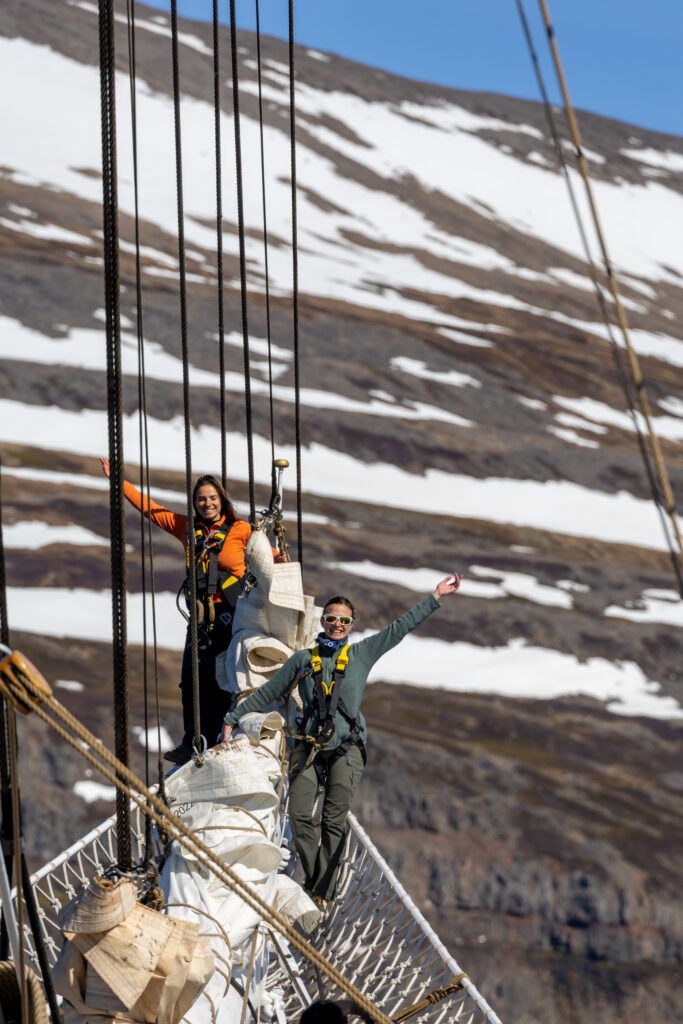 Science students participating in the Ocean Training Course on board the Statsraad Lehmkuhl. (Credit: Ocean Media Lab). Two scientists holding on to the ship's ropes and rails and waving at the camera, with a snowy, rocky coast in the background.