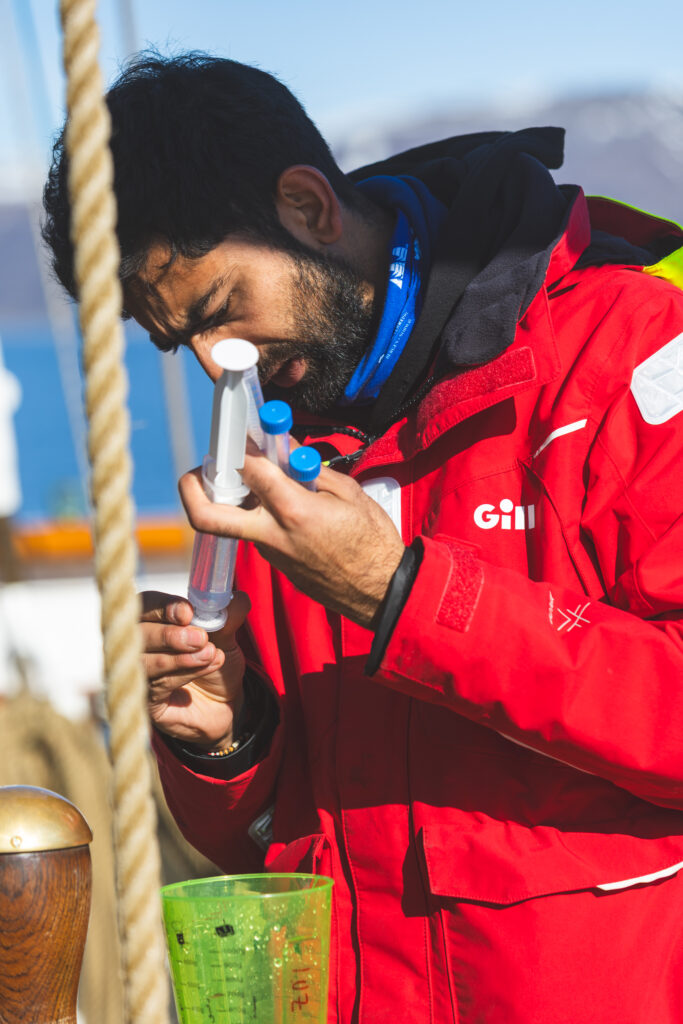 A scientist dress in a red jacket examples the measurement of a large syringe, standing on the deck of a ship.